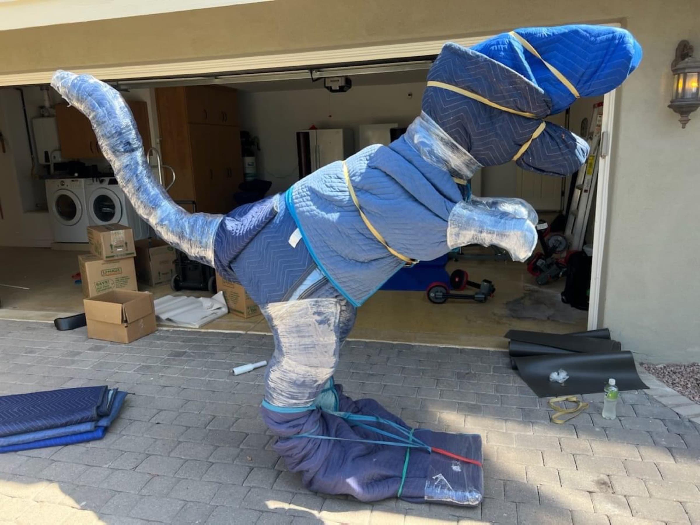 A dinosaur sculpture in a customer's driveway, wrapped in blue quilted moving pads, stretch wrap, and banding, ready to load